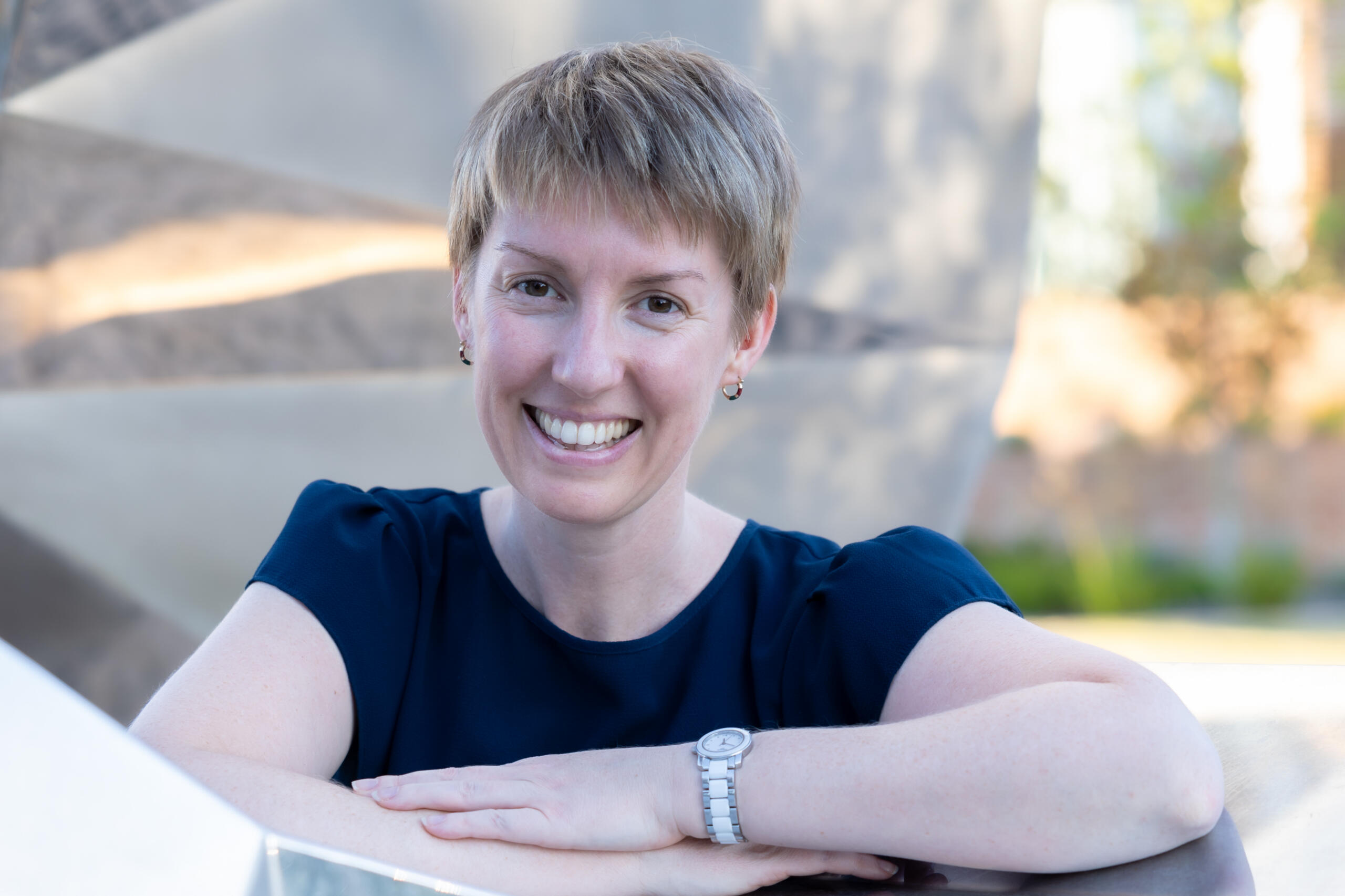 Ria stands behind, and leans against an outdoor sculpure made of metal.